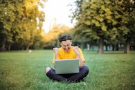一名身穿黄色衣服的年轻人坐在阳光明媚的公园里，用笔记本电脑庆祝成功