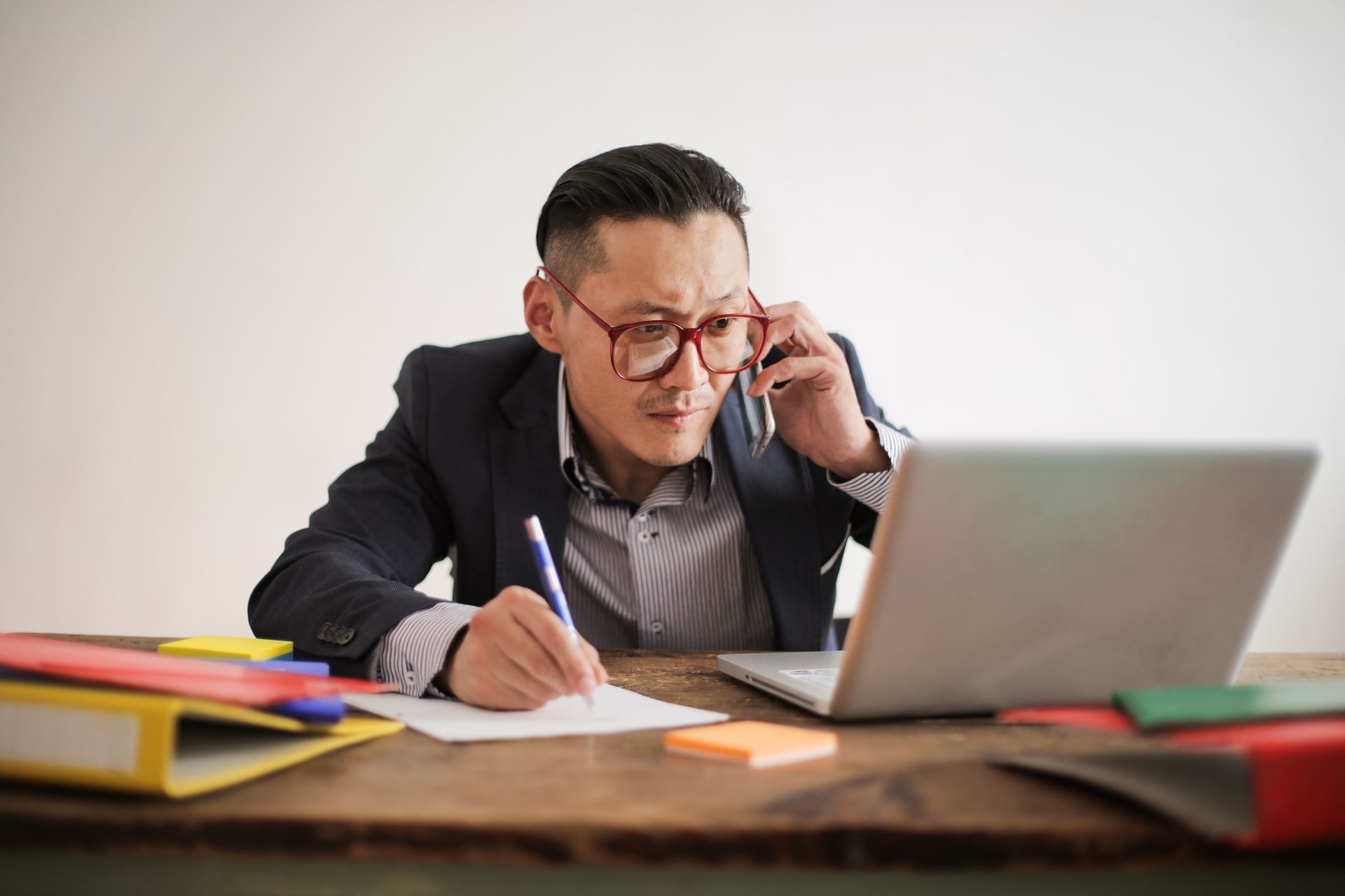 Businessman wearing eyeglasses working intensely on laptop at office desk, with smartphone and documents.