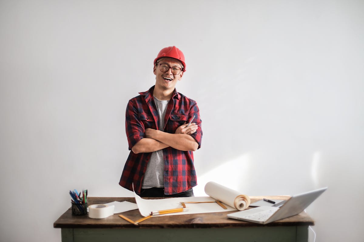 Smiling casual man in hardhat and glasses holding arms crossed looking at camera while standing at desk with paper draft and stationery