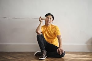 Asian guy in casual clothes using paper cup with thread as telephone while sitting on floor against white wall