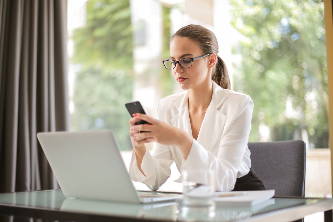 Free Concentrated young woman in formal clothes and eyeglasses sitting at glass table with laptop while surfing smartphone Stock Photo