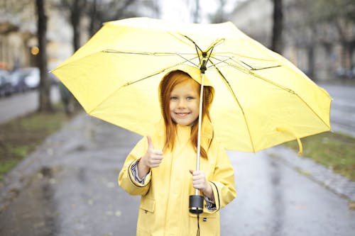 Foto De Niña Sonriente En Impermeable Amarillo Sosteniendo Un Paraguas Amarillo Mientras Da Un Pulgar Hacia Arriba