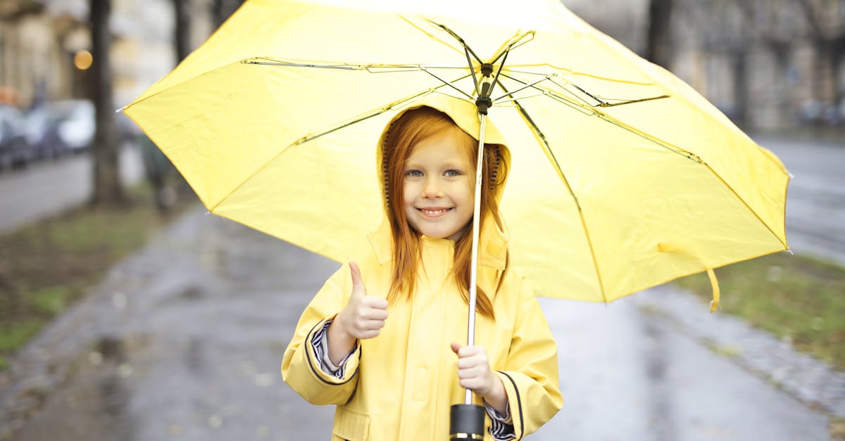 Photo of Smiling Girl in Yellow Raincoat Holding a Yellow Umbrella While Giving One Thumbs Up