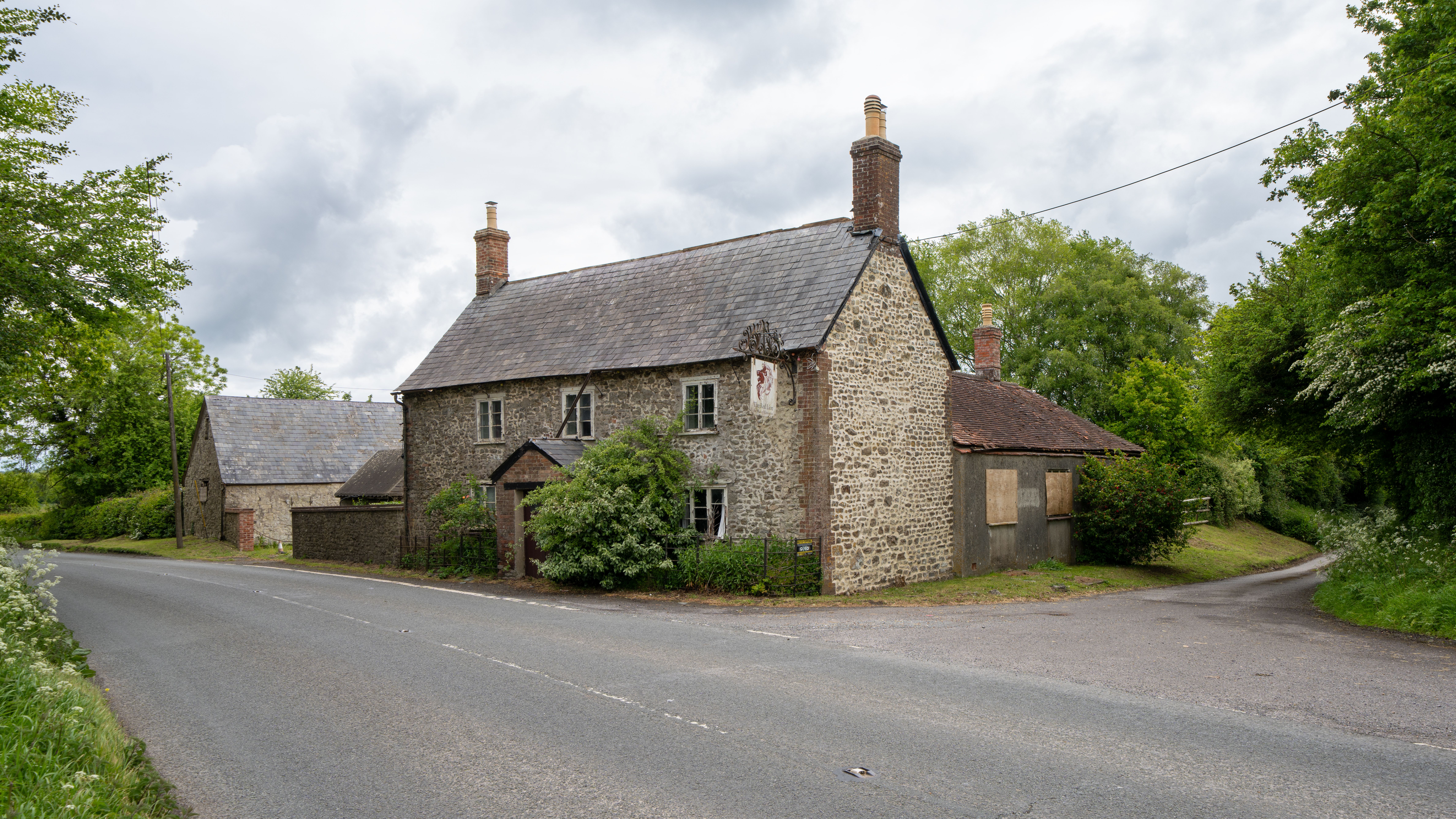 An abandoned stone building resembling a pub with overgrown greenery, set by a quiet rural road.