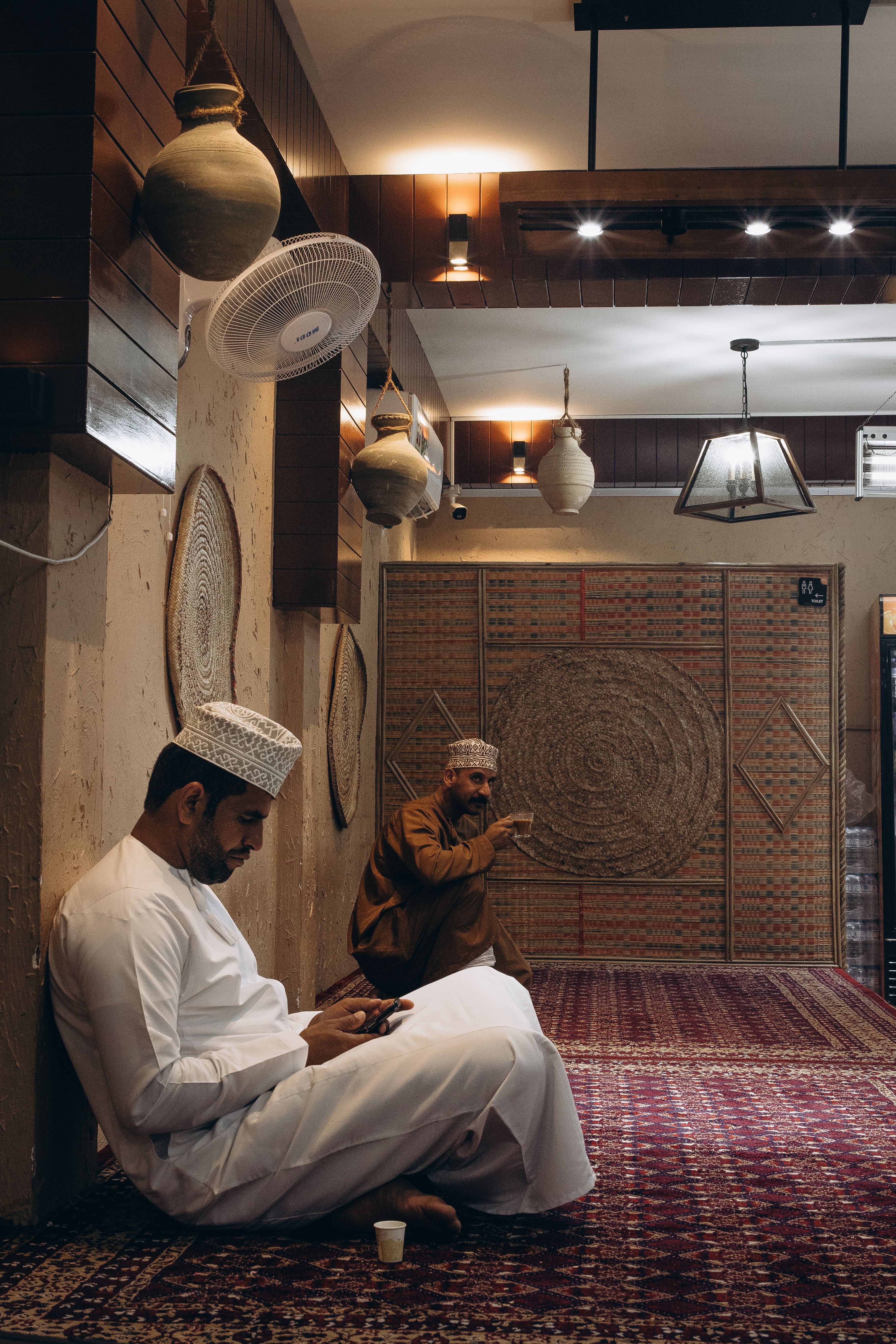 Two men sit indoors on a traditional carpet in Muscat, Oman, featuring ethnic decor.