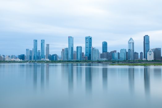 A serene view of modern skyscrapers reflecting on calm water under a cloudy sky.