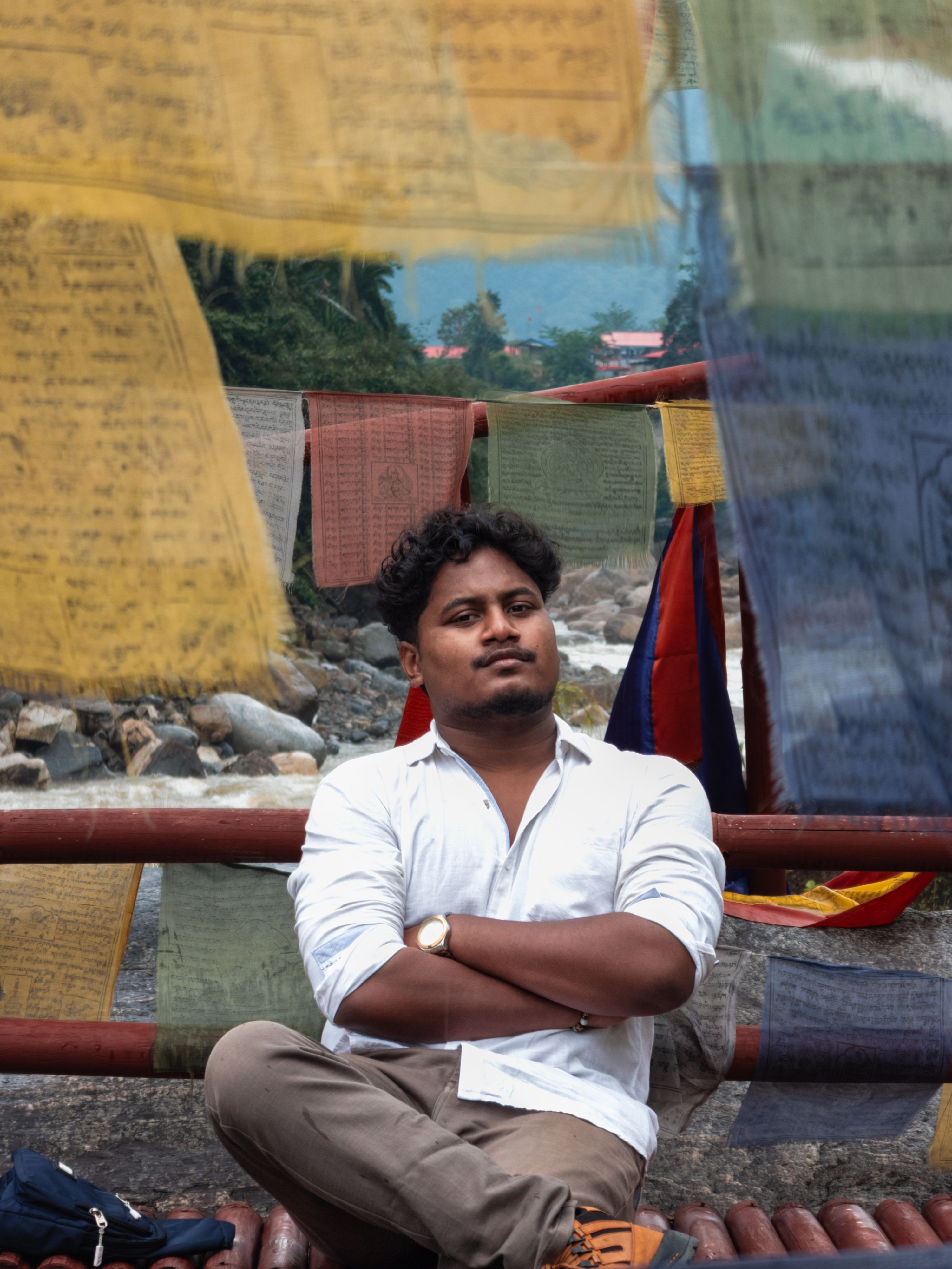 Man sitting calmly near vibrant prayer flags with mountains in the background.