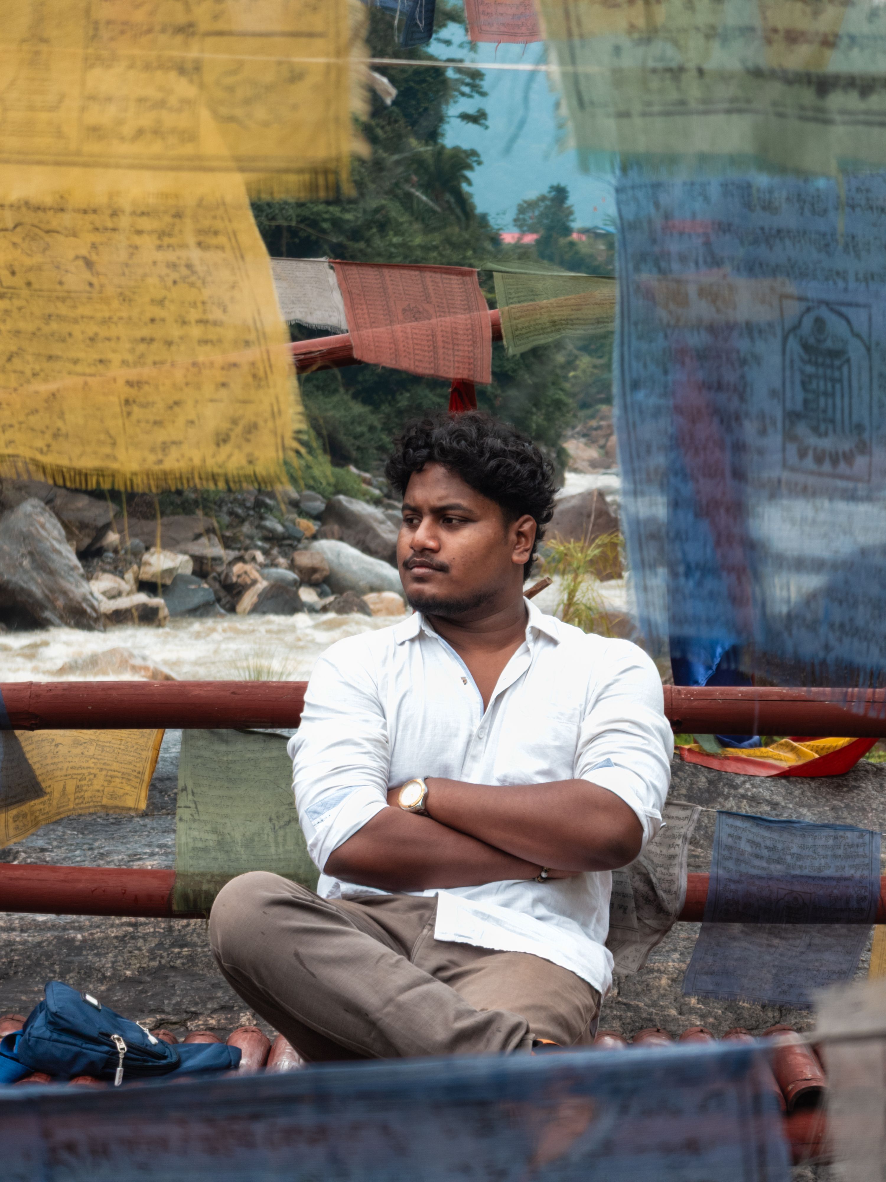 Man sitting contemplatively by a rushing river framed by colorful prayer flags. Tranquil scene.