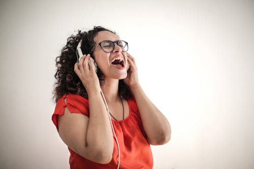 Gratis Foto Wanita Bernyanyi Dengan Kacamata Merah Atasan Dan Bingkai Hitam Mendengarkan Musik Di Headphone Foto Stok