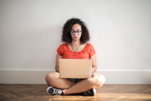 橙色乘員脖子t卹坐在棕色木地板上使用筆記本電腦的女人
