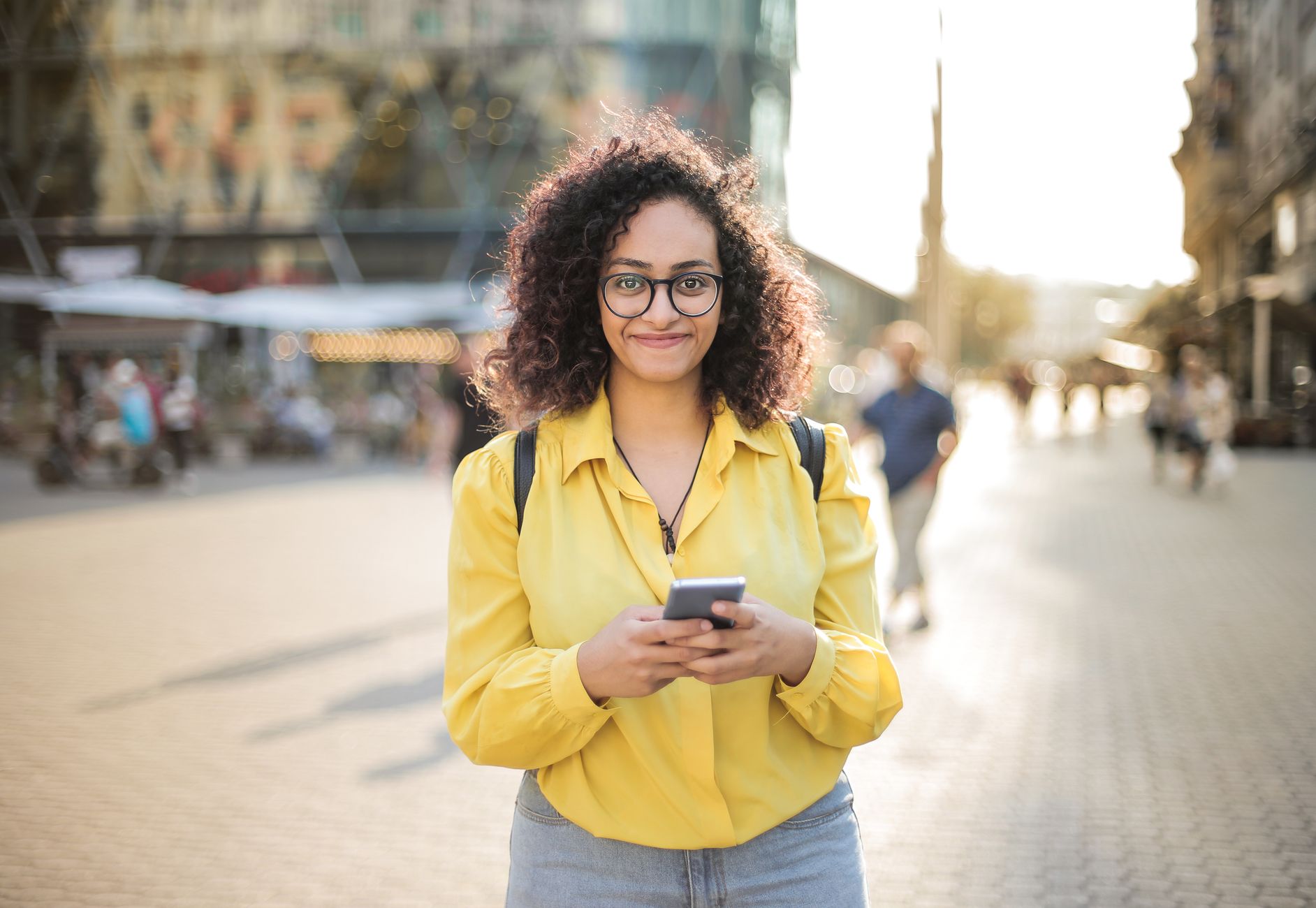 Cheerful woman with curly hair and glasses using smartphone outdoors in a sunny urban setting.