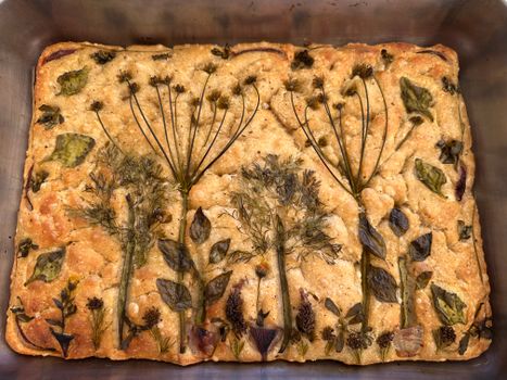 Artistic focaccia bread topped with herbs and flowers in a baking tray, presenting a natural design.
