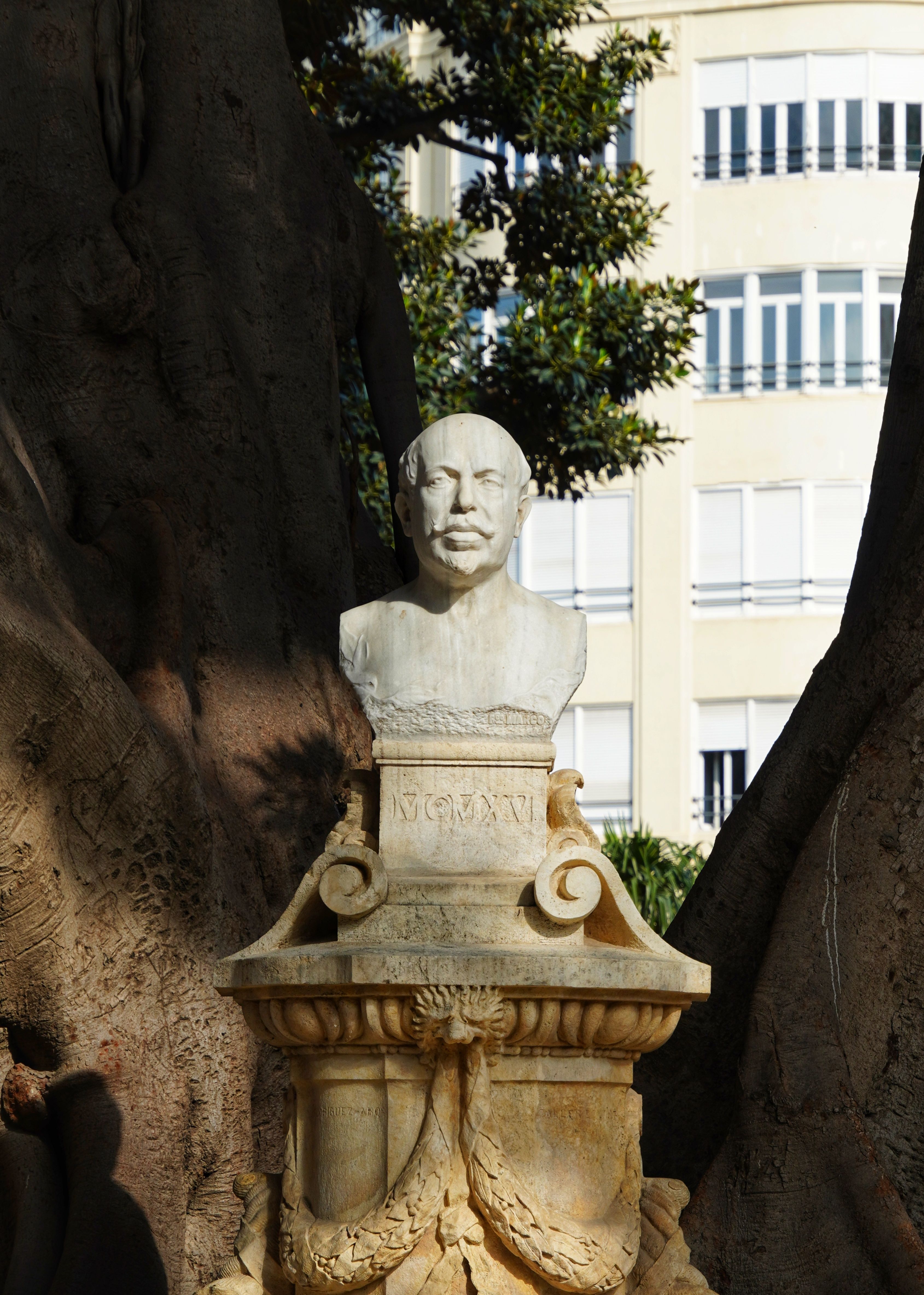 A stone bust on a pedestal outdoors surrounded by trees and urban buildings.