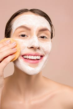 A woman smiling while applying a facial mask using a sponge for skincare.