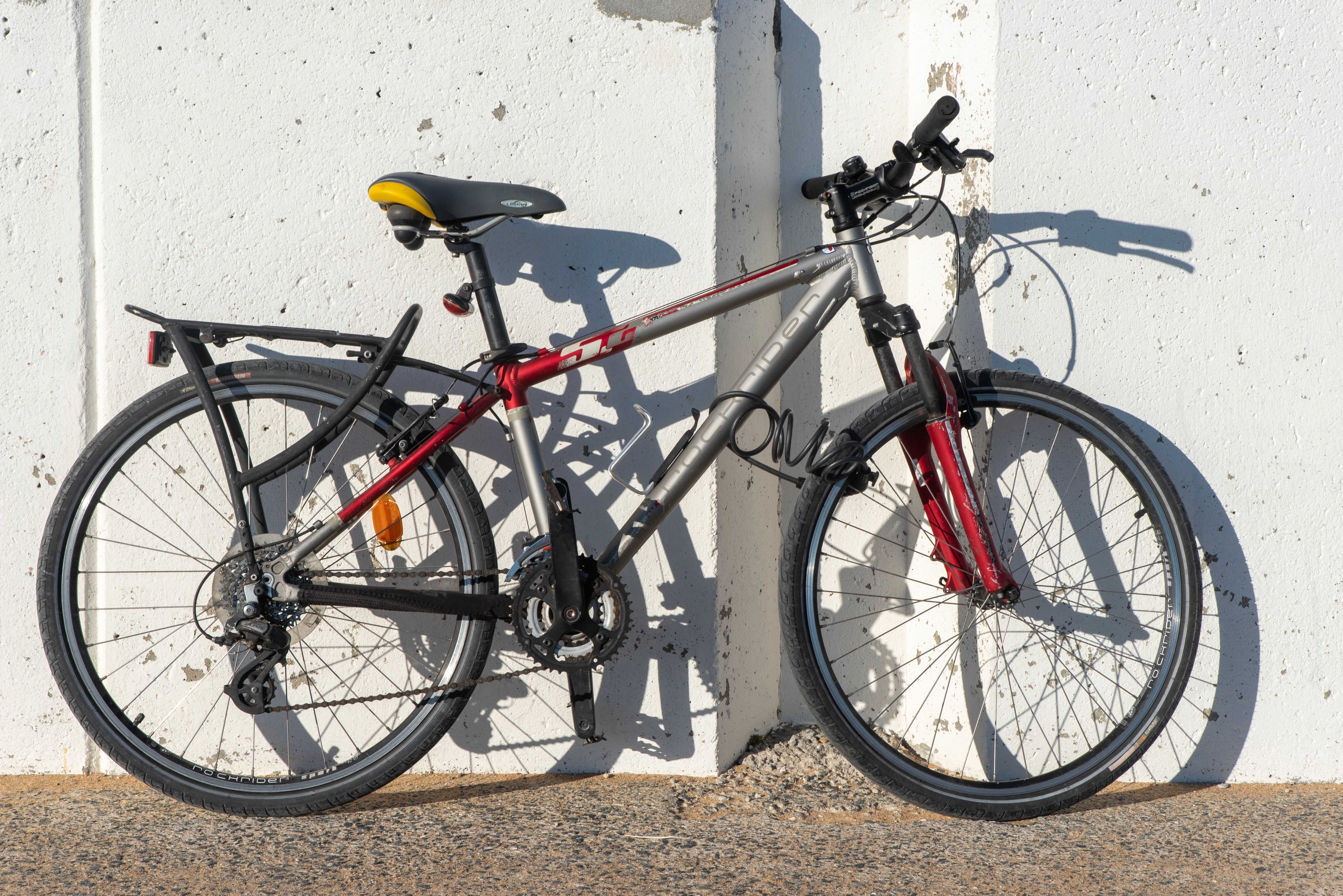 A bicycle with a red and gray frame is parked against a sunny concrete wall.