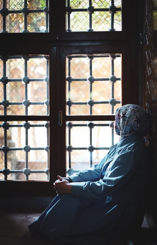 Free A woman in a blue dress and headscarf sits by a large, ornate window, deep in thought. Stock Photo