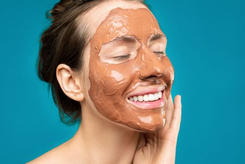 Free Close-up of a woman smiling with a clay mud mask on her face on a turquoise background. Stock Photo