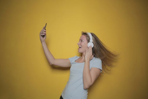 Foto Tampak Samping Wanita Tersenyum Dengan Tank Top Putih Mendengarkan Musik Di Headphone Saat Mengambil Selfie