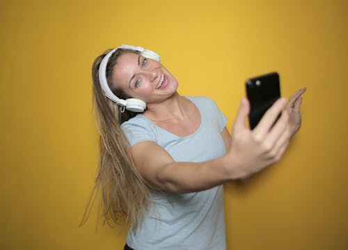 Foto De Uma Mulher Sorridente, Vestindo Uma Camiseta Branca, Ouvindo Música Nos Fones De Ouvido Enquanto Tira Uma Selfie