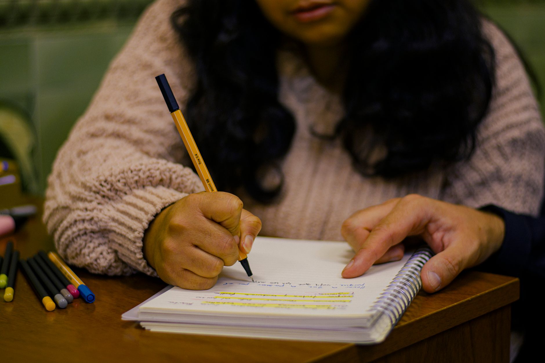 Close-up of a student writing in a journal with colored pens on a wooden desk.