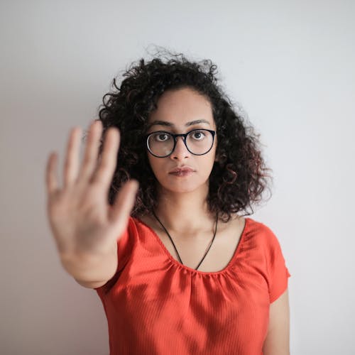Портретное фото женщины в красном топе в очках в черной оправе, протягивающей руку в жесте остановки