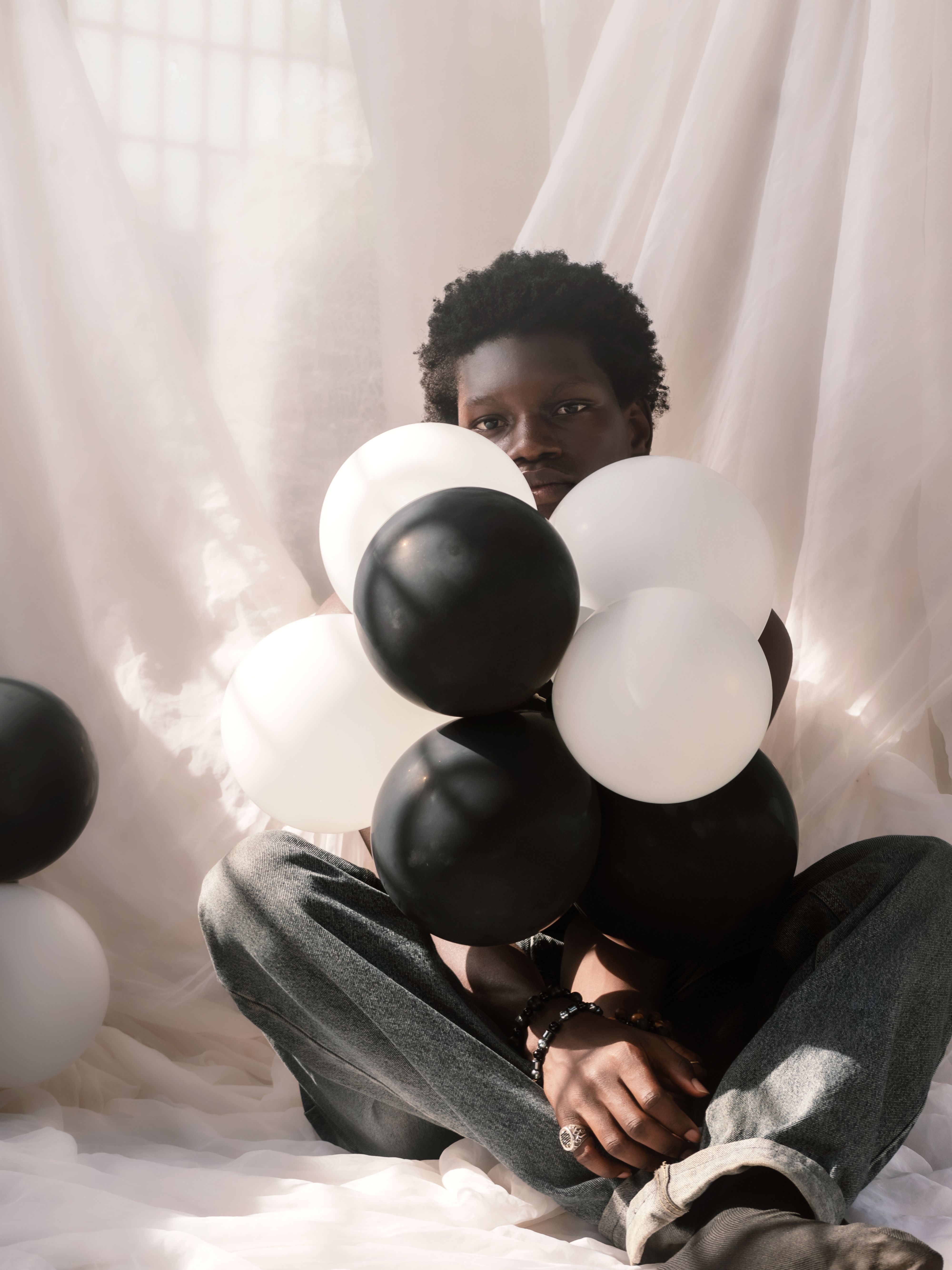 A young adult sits surrounded by black and white balloons in soft lighting.