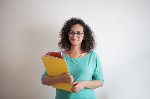 Portrait Photo of Smiling Woman in Teal Top Carrying Folders While Standing In Front of White Background