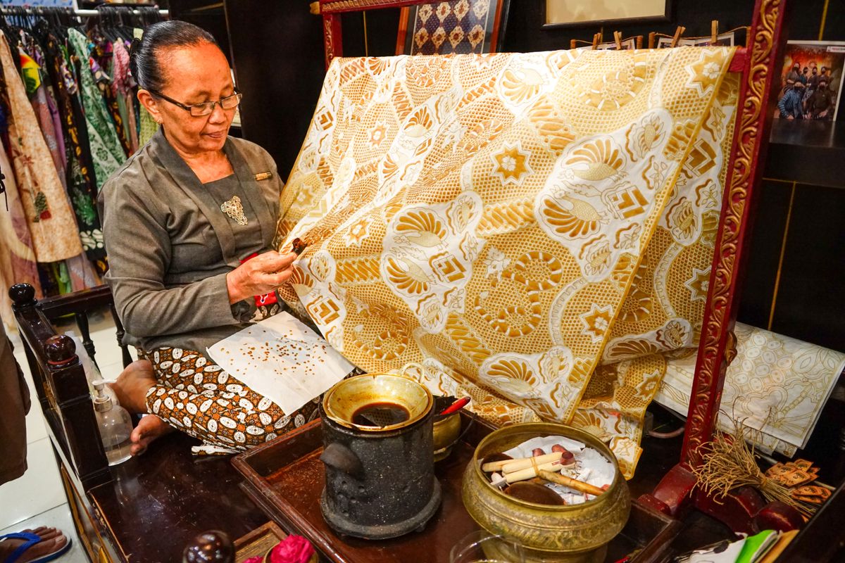 A craftswoman seated at a wooden frame with a length of cream and gold batik cloth stretched taut, wax pot and tools on the table beside her.
