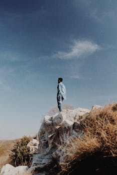 一名男子独自站在蔚蓝天空下的岩石悬崖上，象征着孤独与冒险