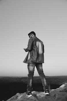 Monochrome image of a young man using a smartphone on a rocky outdoor landscape.