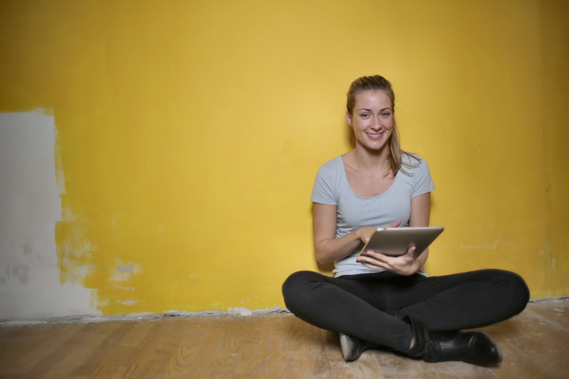 A woman enjoying technology while seated indoors, smiling at a tablet against a vibrant yellow wall.