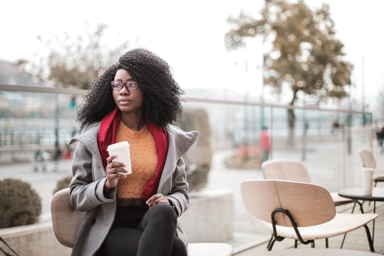 Selective Focus Photo Of Woman In Gray Coat Sitting On Wooden Chair Holding A To Go Cup Of Coffee