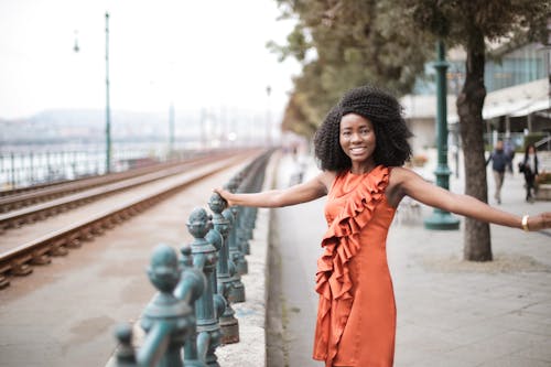 Selective Focus Photo of Smiling Woman in Orange Sleeveless Dress Posing By Metal Railing