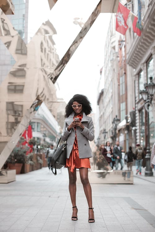 Photo of Woman in Gray Coat and Orange Dress Standing Alone While Using Her Phone