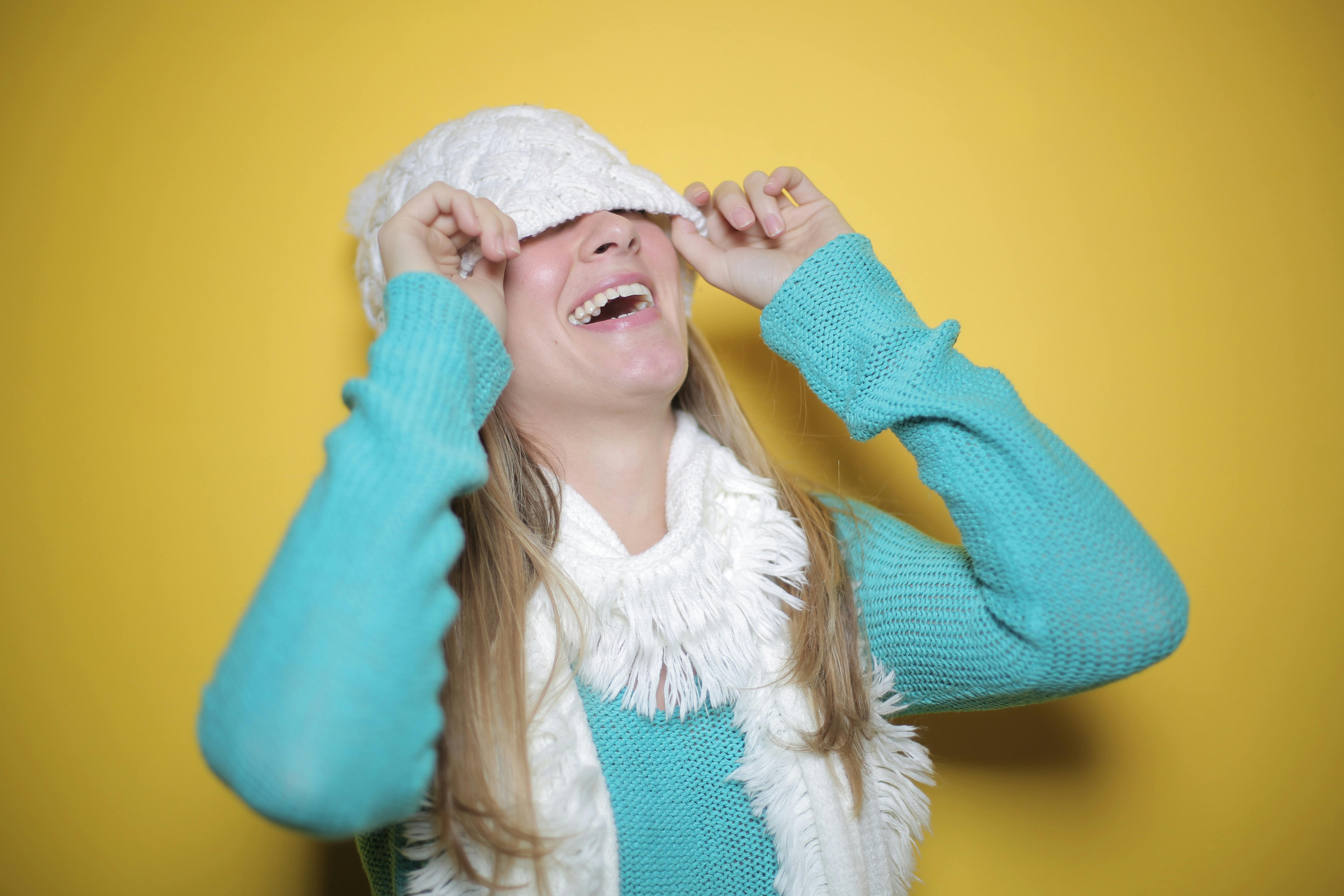 一位快乐的女子穿着蓝色毛衣、戴着围巾，在亮黄色背景下调整无檐帽时顽皮地笑着