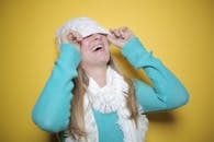 一位快乐的女子穿着蓝色毛衣、戴着围巾，在亮黄色背景下调整无檐帽时顽皮地笑着