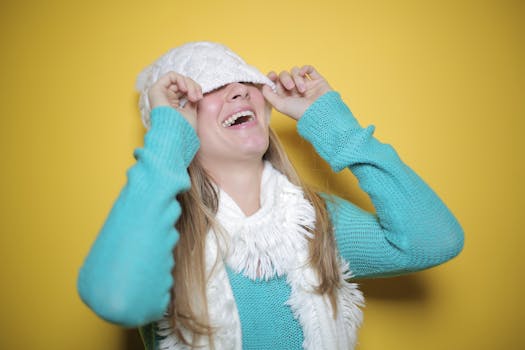 一位快乐的女子穿着蓝色毛衣、戴着围巾，在亮黄色背景下调整无檐帽时顽皮地笑着