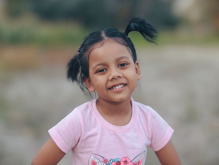 A cheerful young girl with pigtails smiles at the camera while standing outdoors.
