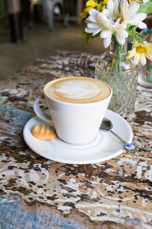 Free A beautifully crafted latte on a rustic table with flowers and a biscuit, perfect for a cozy cafe atmosphere. Stock Photo