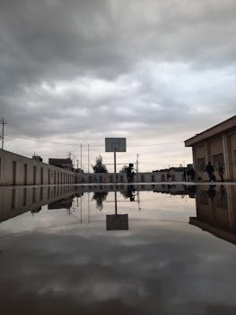 Cloudy sky reflecting in water of huge puddle on basketball playground located on territory of school
