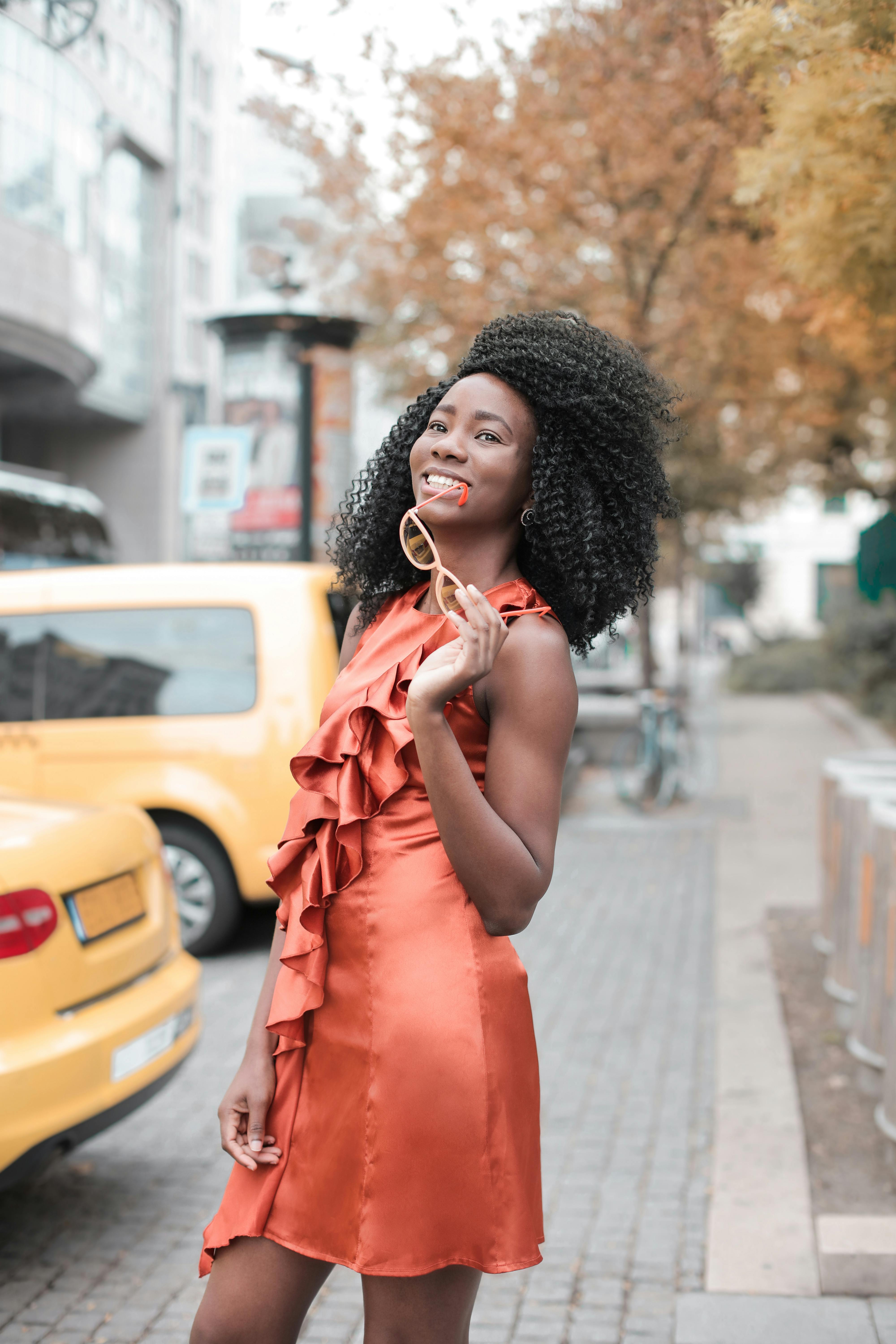 一位身穿橙色连衣裙的时尚女子，在黄色汽车旁的城市人行道上微笑