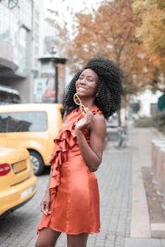 一位身穿橙色连衣裙的时尚女子，在黄色汽车旁的城市人行道上微笑