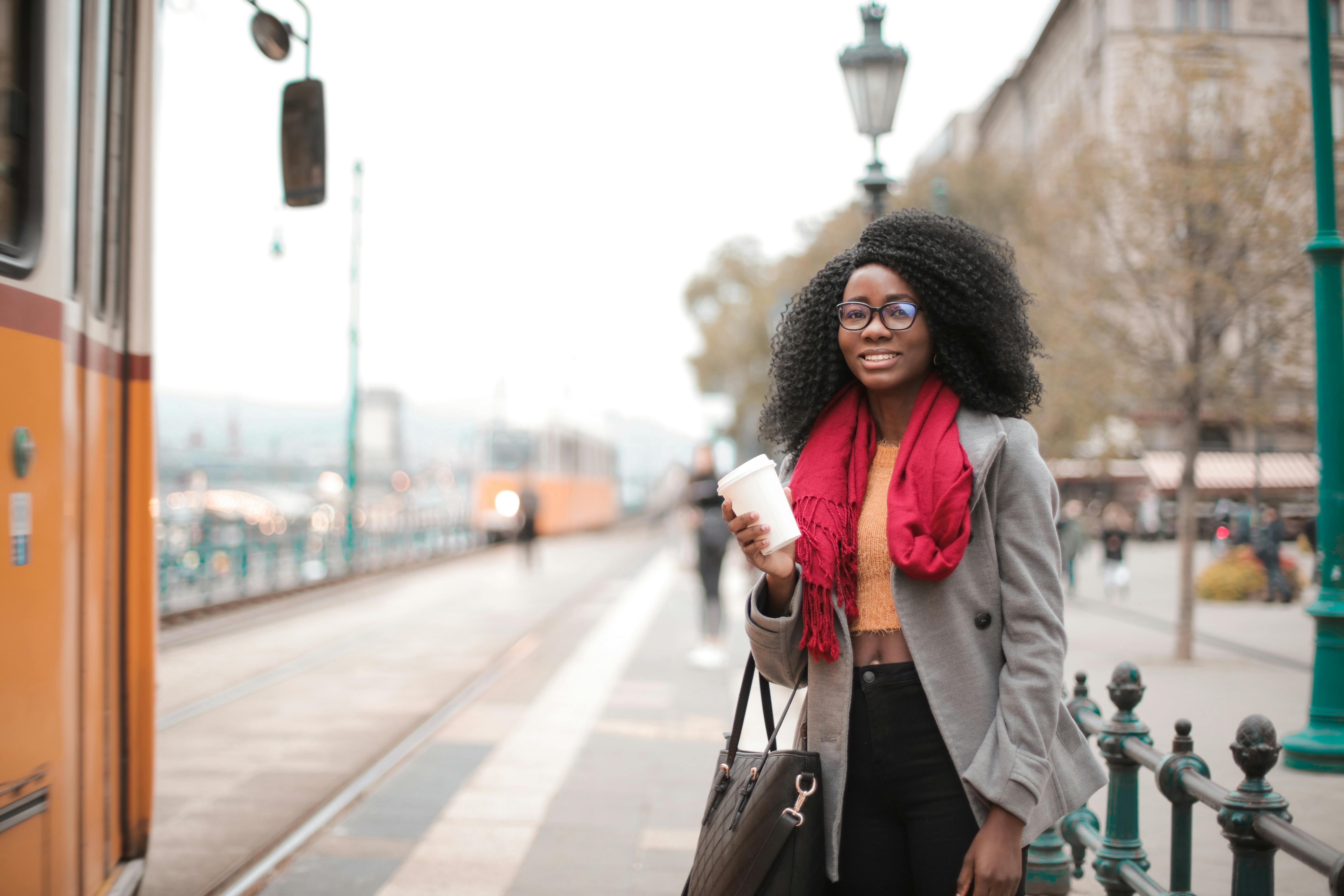 一位穿着休闲装的女子微笑着，端着咖啡在城市车站等电车