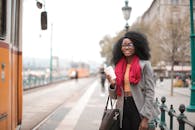 一位穿着休闲装的女子微笑着，端着咖啡在城市车站等电车