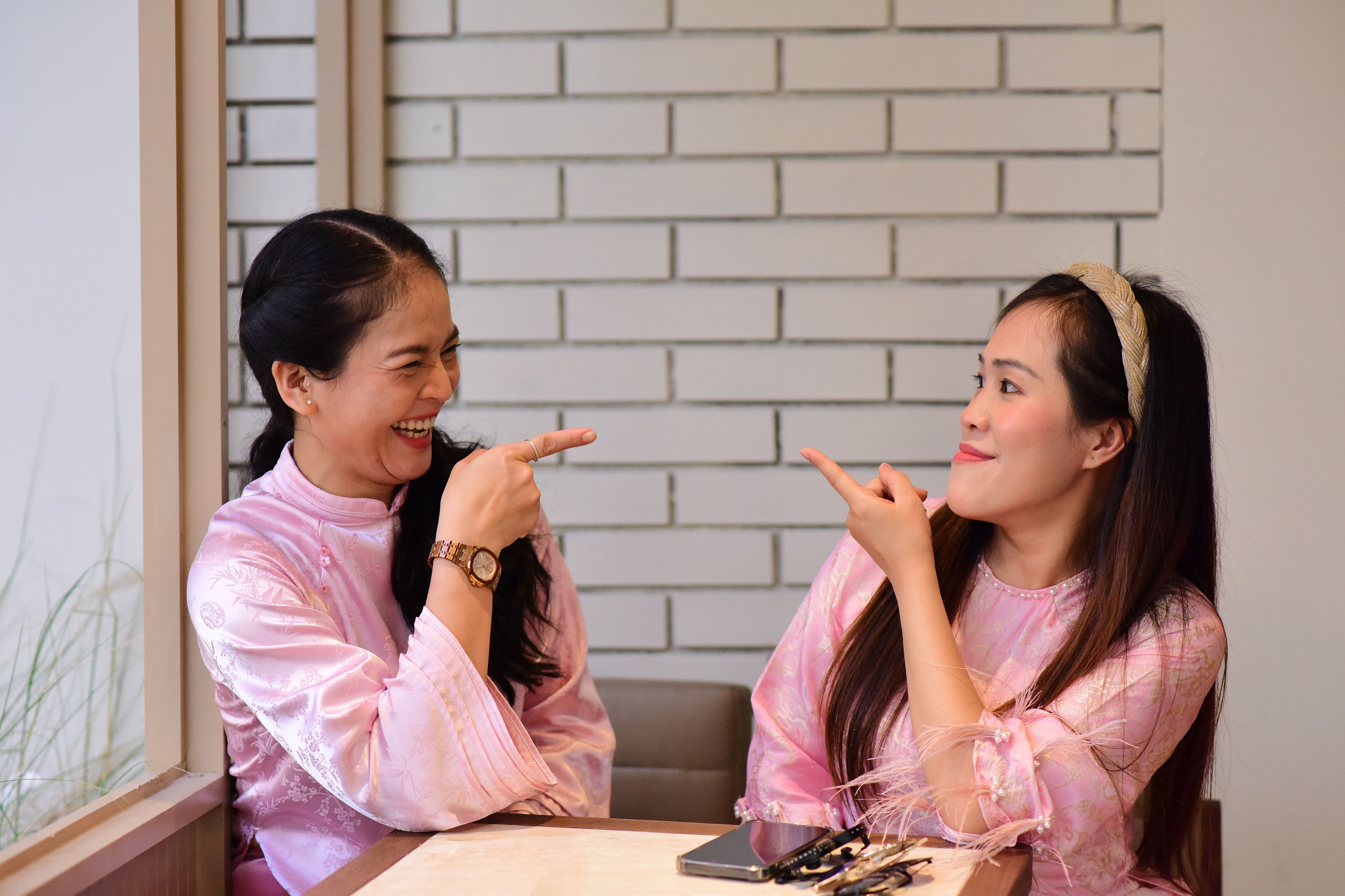 Two women joyfully pointing at each other while laughing in a cozy indoor setting.