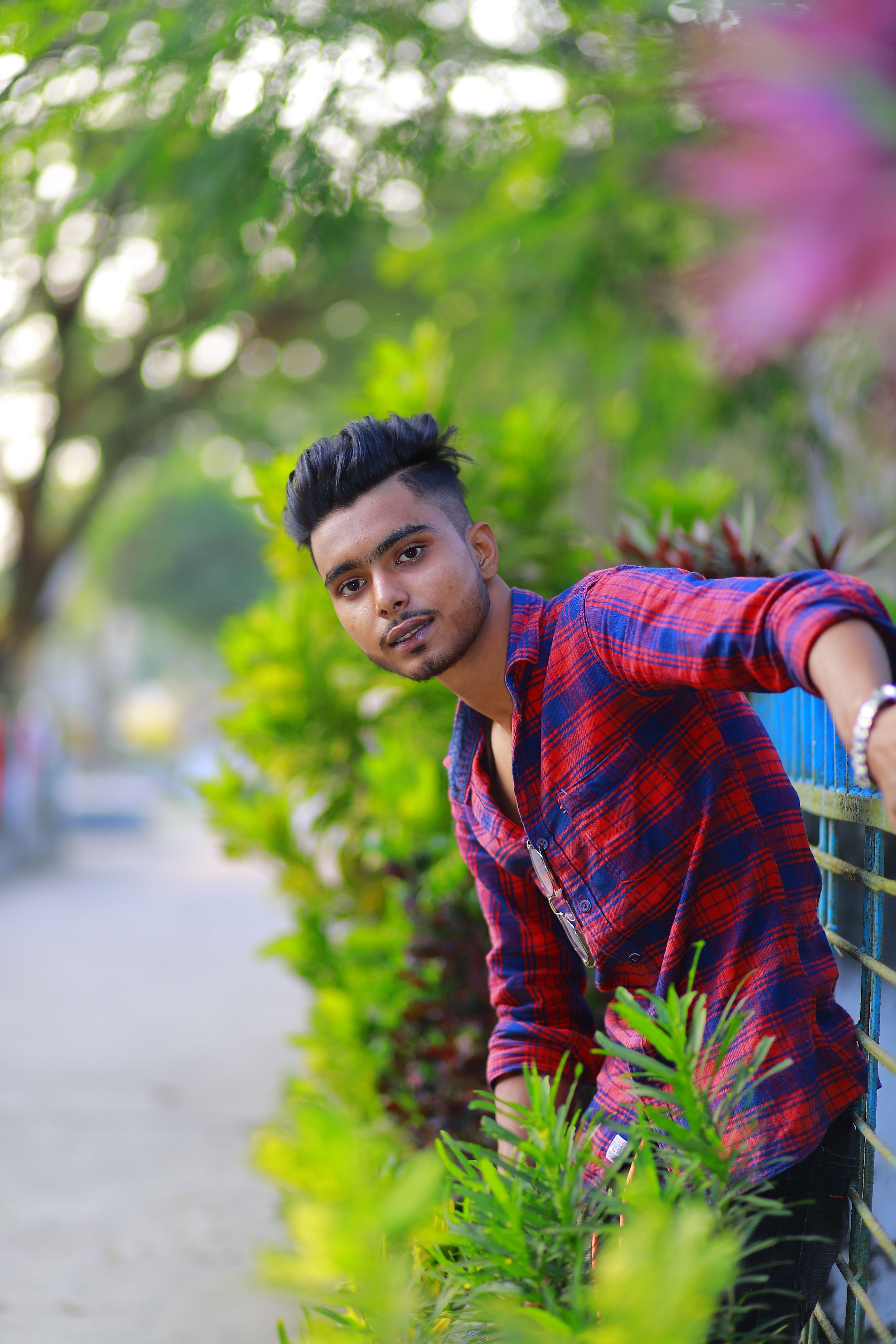 Casual portrait of a young man outdoors, wearing a red plaid shirt, surrounded by greenery.