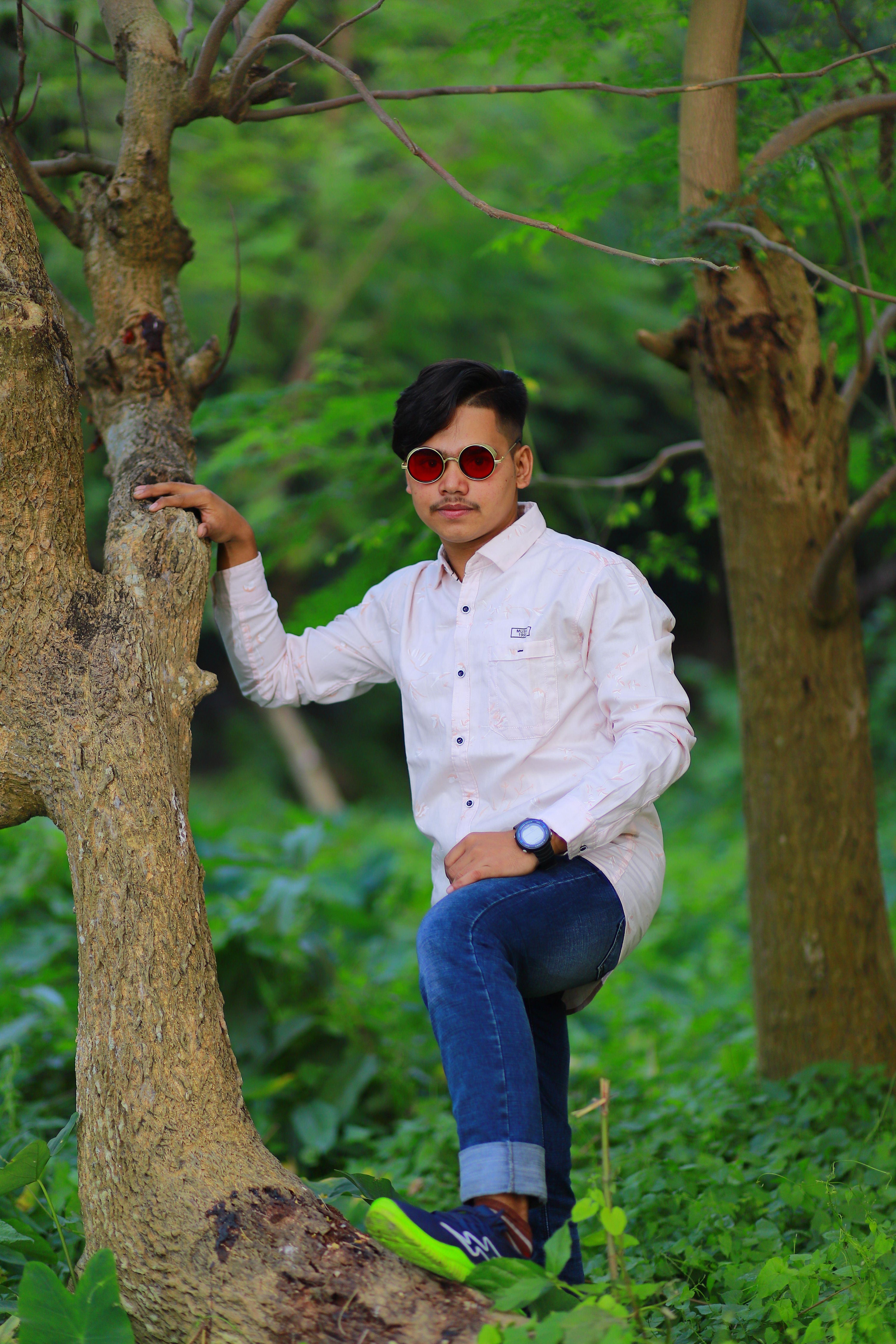 Stylish young man in a white shirt and jeans posing confidently against a tree in a green forest.