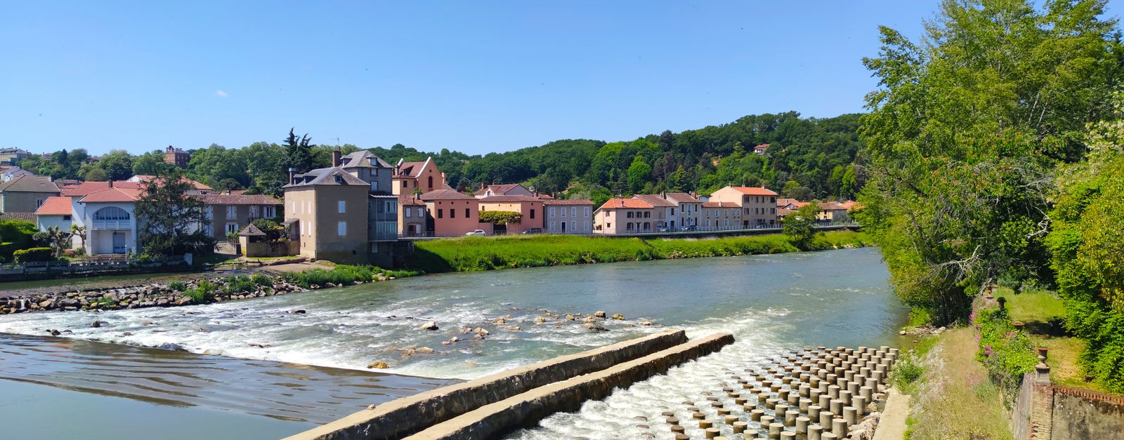A scenic view of Aire-sur-l'Adour with colorful houses along the river under a clear blue sky.
