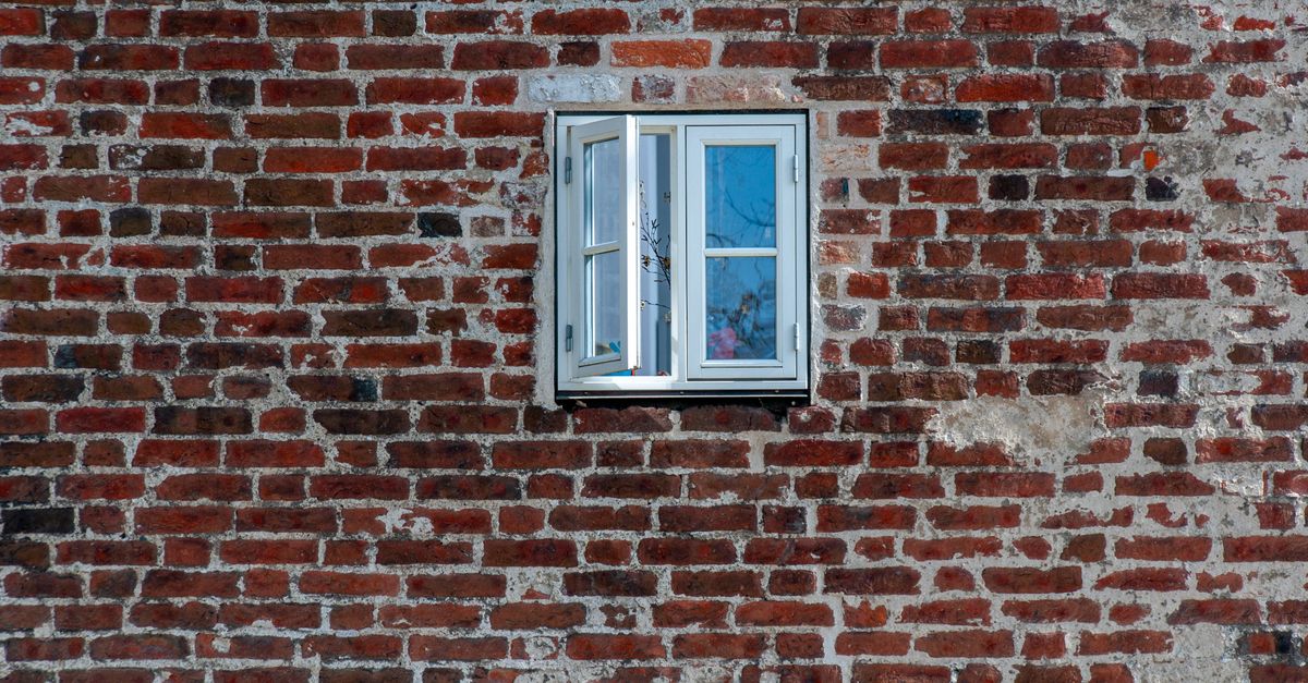 Geöffnetes weißes Fenster in einer alten roten Backsteinwand, Symbol für Lüftung und Feuchtigkeit im Altbau.