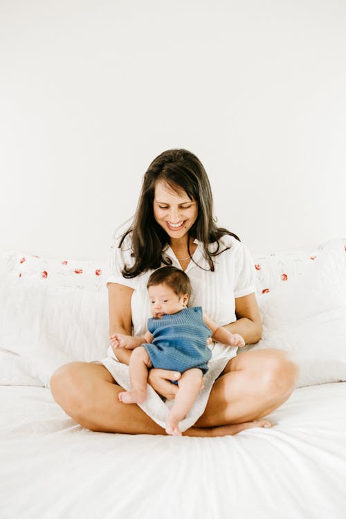 Free Photo of a Smiling Woman Carrying Her Baby While Sitting on a Bed Stock Photo